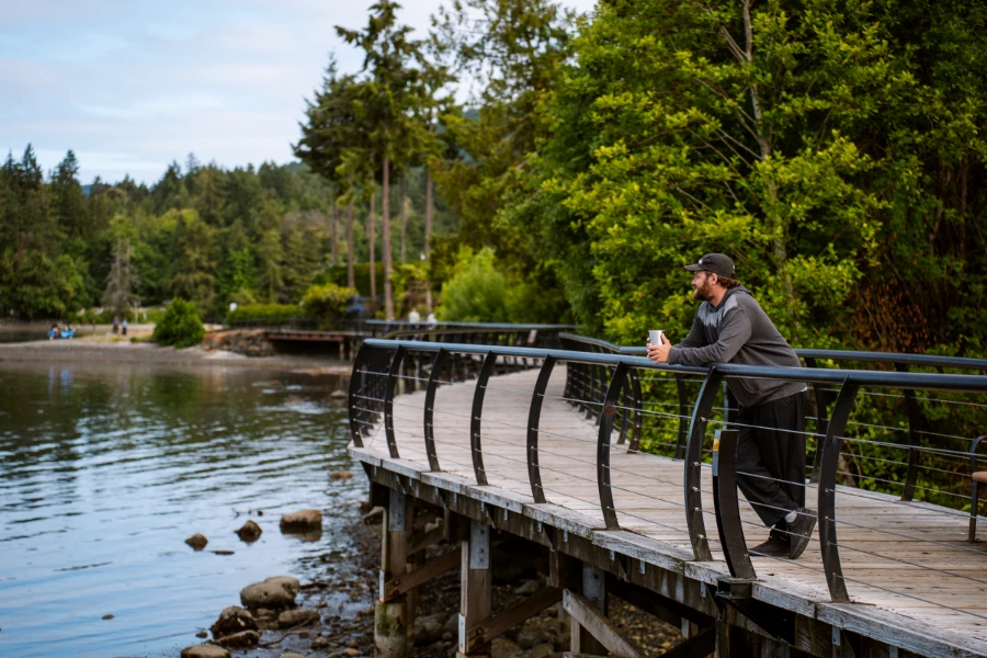 A man leaning on a metal railing that lines a flat wooden boardwalk along the shore of the Salish Sea