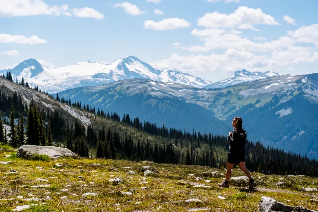 A woman walking along a dirt path in the Blackcomb Alpine with Whistler Mountain visible across the valley