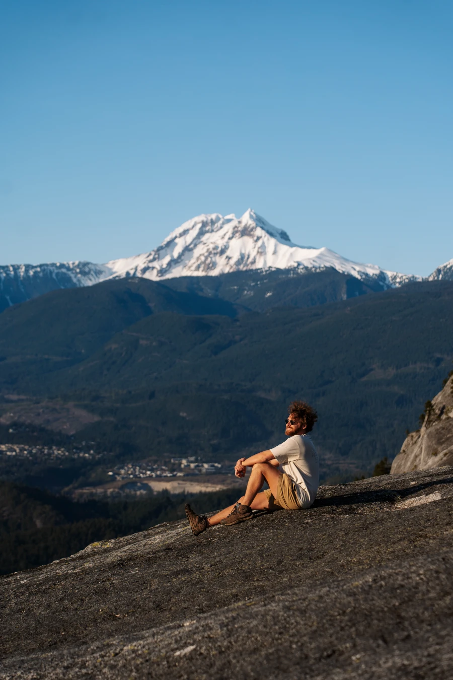 A man sitting on the bare stone peak of the Stawamus Chief with Mount Garibaldi towering over the valley in the distance