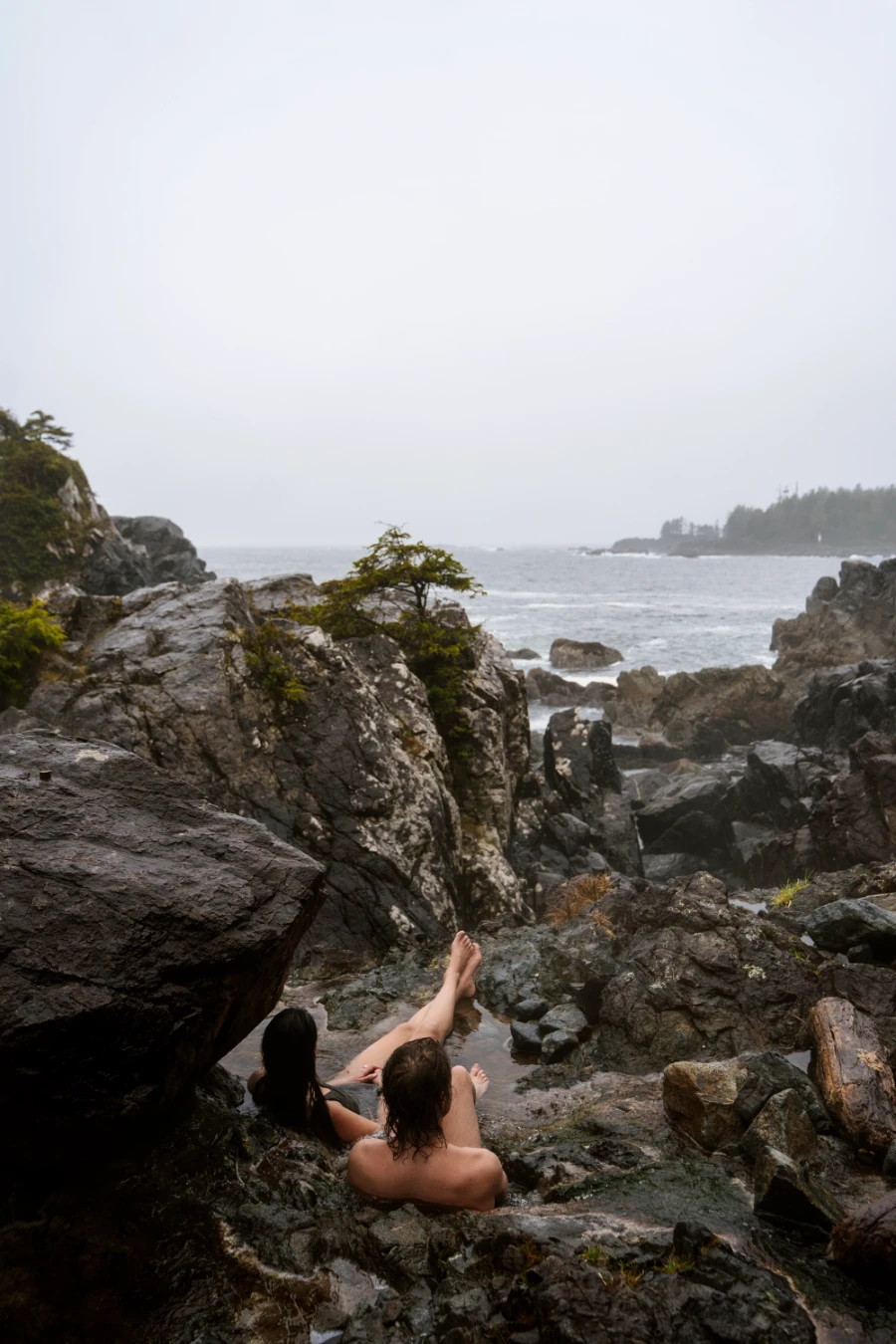 A man and a woman sit in a small rocky pool of water looking out to the ocean at Hot Springs Cover near Tofino, BC