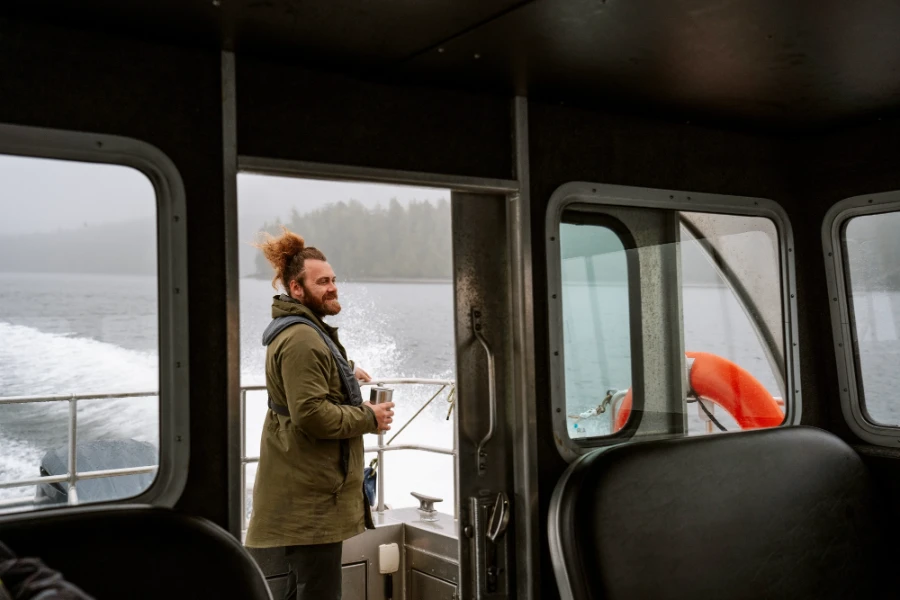 Man stands at the back of a moving boat with a coffee in his hand looking out to a misty rainforest on the shore