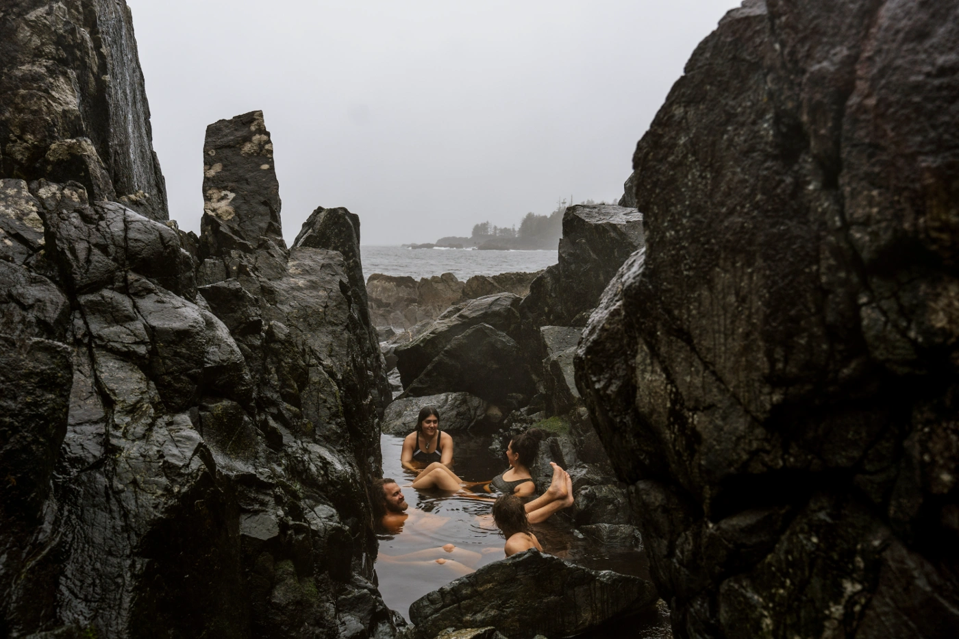 Four people sit laughing and chatting in a shallow rocky pool of water at Hot Springs Cover near Tofino, BC