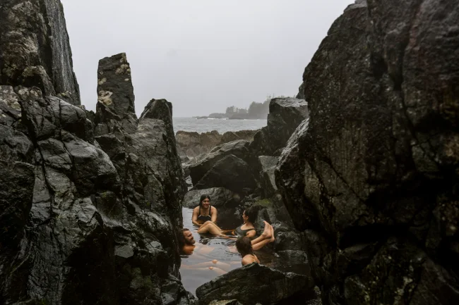 Four people sit laughing and chatting in a shallow rocky pool of water at Hot Springs Cover near Tofino, BC