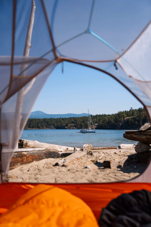 An anchored sailboat just off the coast of Thormanby Island framed by the entrance flap of a tent that is set up on the beach