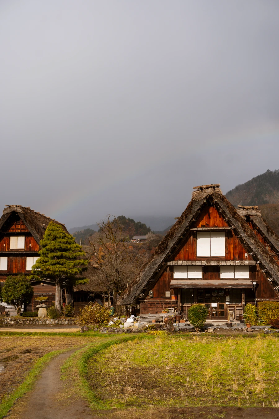 A dirt path leads to multiple traditional Japanese Houes with a rainbow streaking across the sky above it