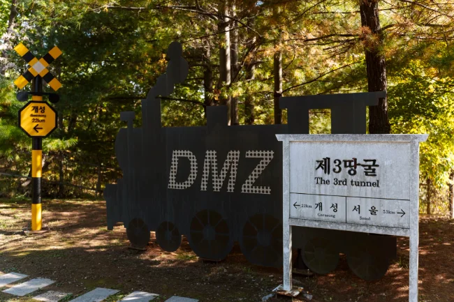 A sign read 3rd tunnel in front of a cut out of an old train with writing on it that reads DMZ