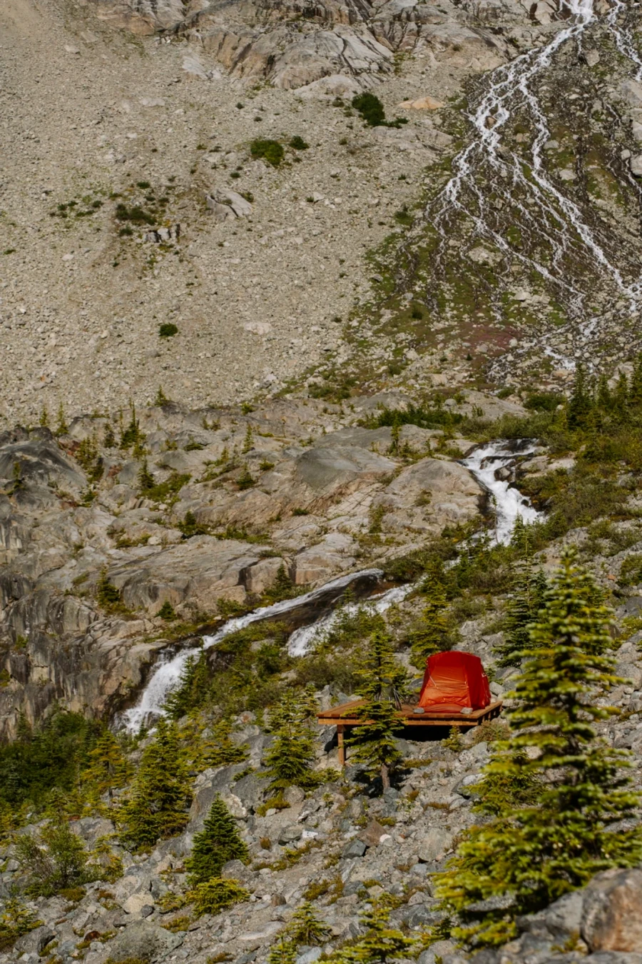 A red tent is set up on a wooden platform with a waterfall cascading down beside it fed by a large glacier