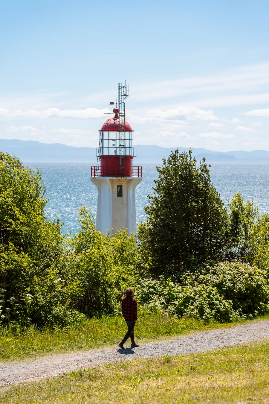 A man walks along a narrow gravel path in front of a white and red lighthouse in Juan De Fuca