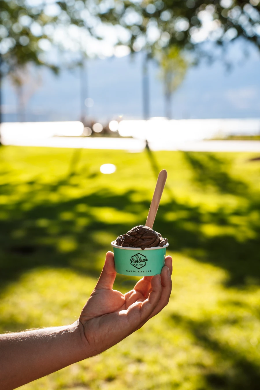A hand hold up a small cup of chocolate ice cream in front of the manicured grass of City Park in Kelowna