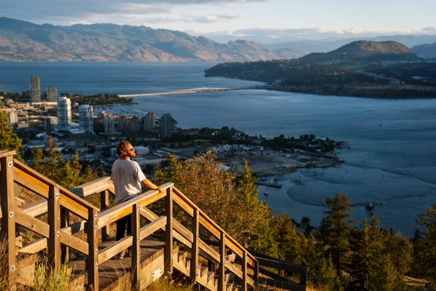 A man stands on a wooden staircase near the top of Knox Mountain, looking down at the cityscape and the adjacent lake from high above Kelowna