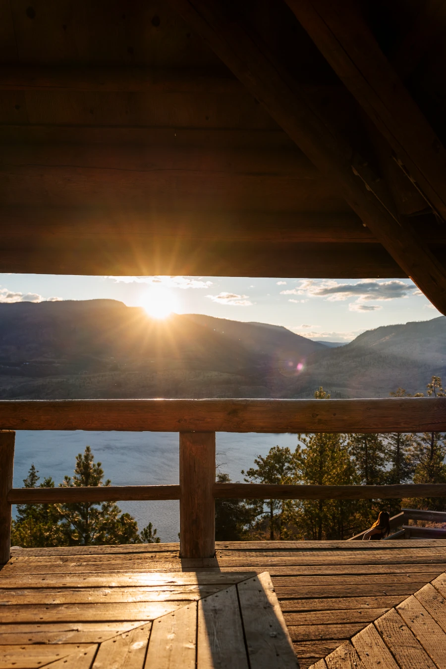 Sunflare shoots in all directions as the sun tucks behind a distant mountain across Okanagan Lake and streams between the sloped ceiling and large railing of a gazebo built on top of Knox Mountain