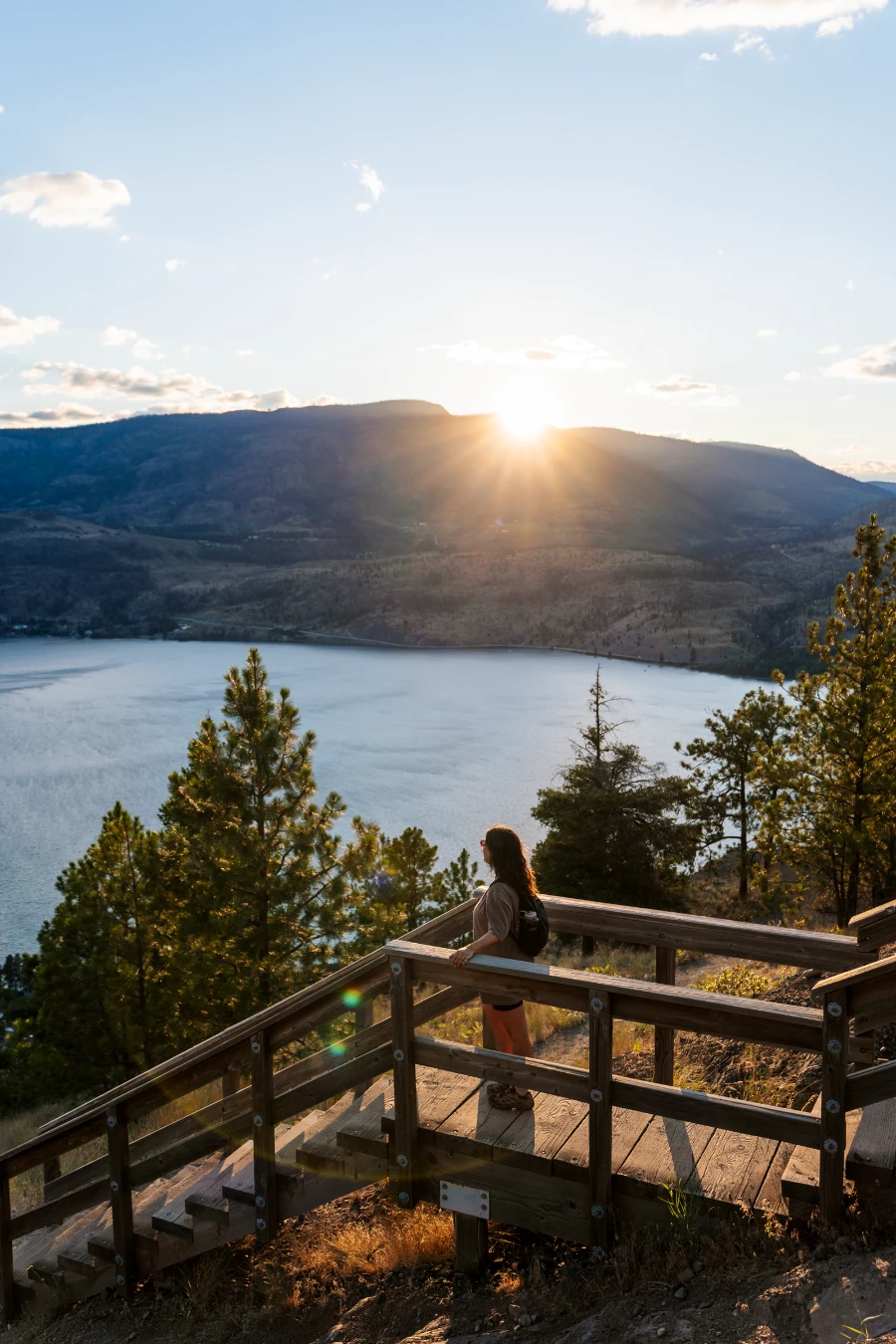 A woman stands on a large wooden staircase that is built at the top of Knox Mountain while the sun flares as it sets below distant mountains across Okanagan Lake