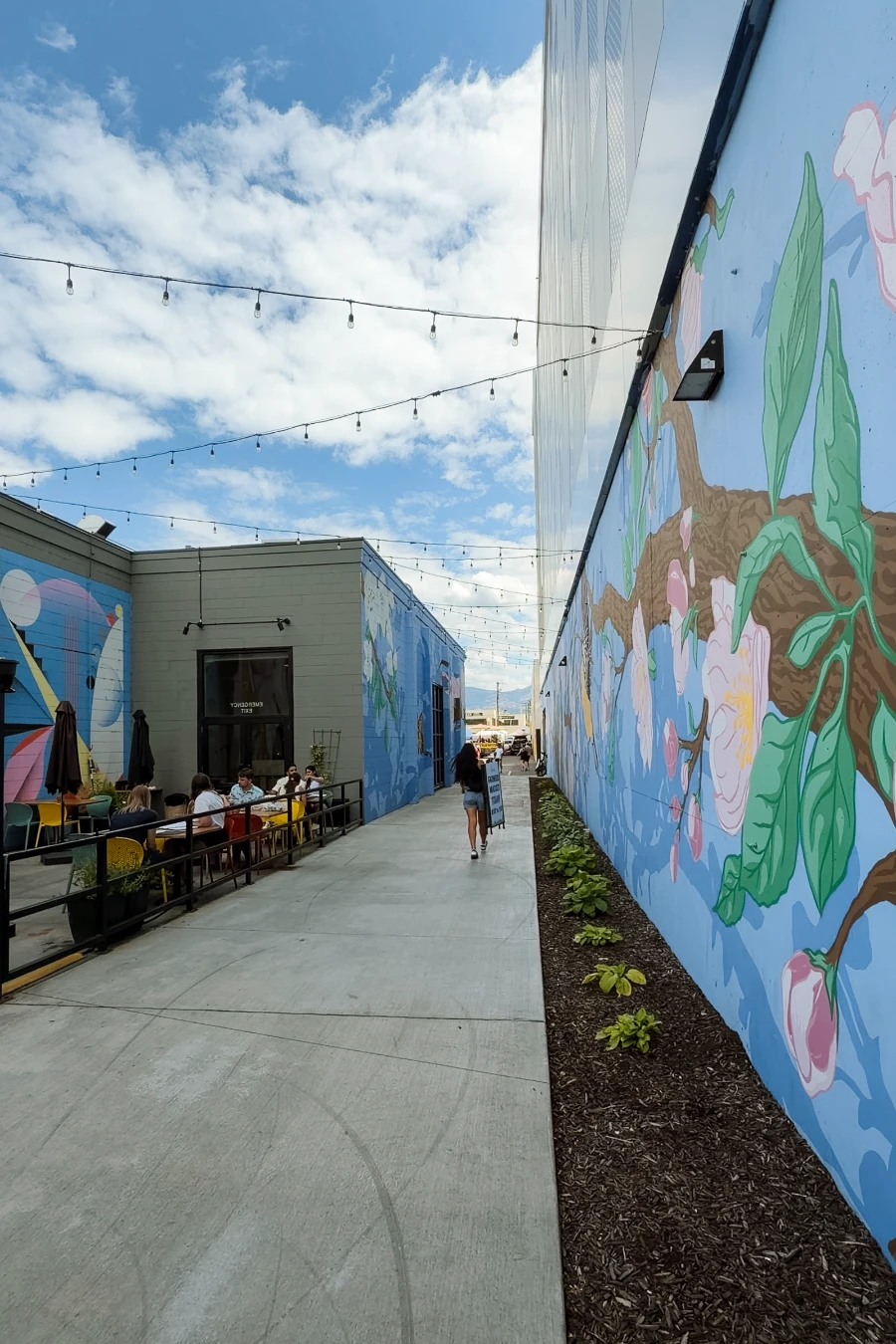 A cement pathway passes by two buildings, with the one on the right painted with a large mural and the one on the left being home to a small patio with more murals painted on the walls behind