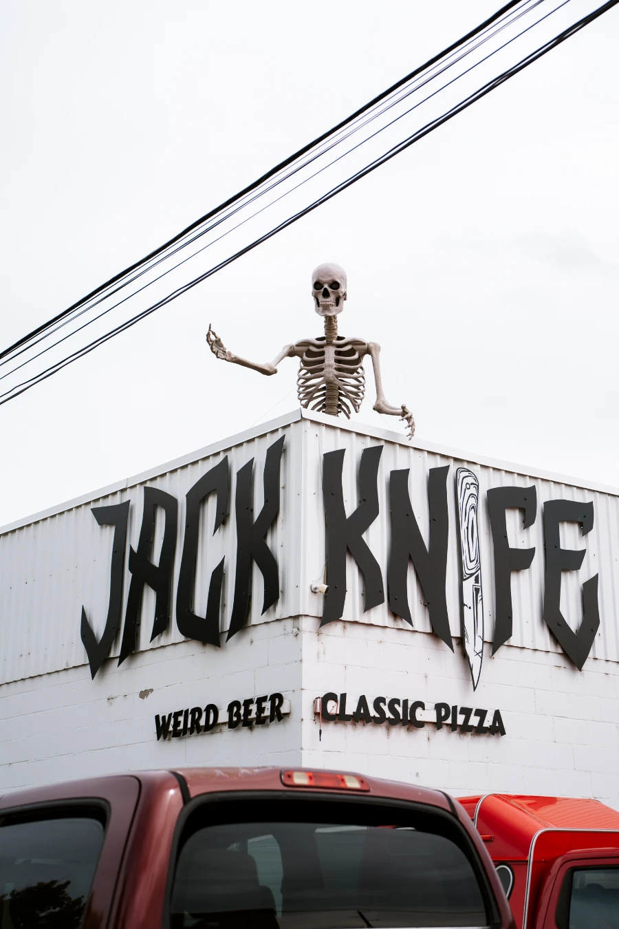 A massive plastic skeleton stands on the roof of a white building with a sign hung on the face of the building reading ‘Jack Knife Weird Beer Classic Pizza’