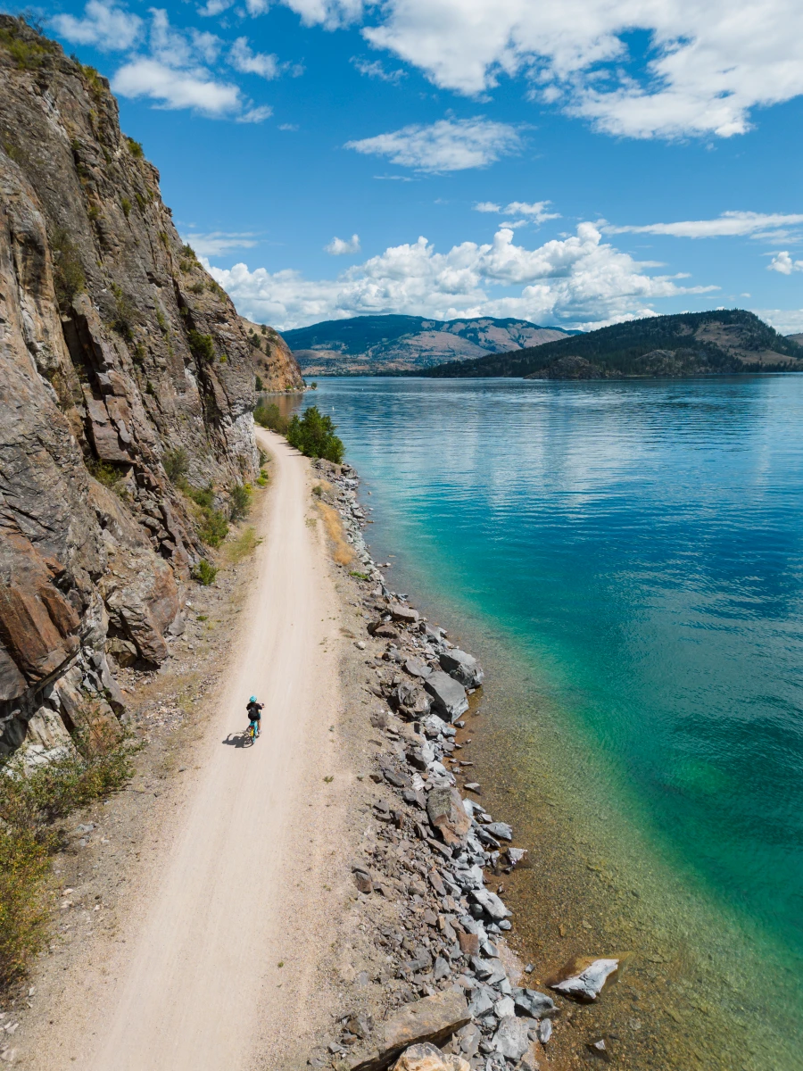 A cyclist rides down the centre of a wide dirt path with a steep stone cliff rising to their left, and turquoise waters to their right