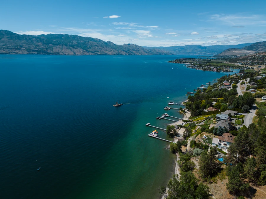 Several homes on the shores of Okanagan Lake with long docks stretching out into the calm water while a single large boat traverses the lake leaving a choppy wake behind it