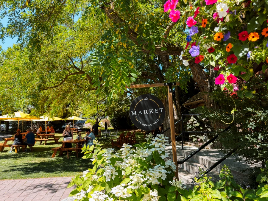 Lush flowers and bushes surround a wooden sign that reads ‘Market’ in front of a well-maintained lawn with several picnic tables that each sport a large yellow umbrella