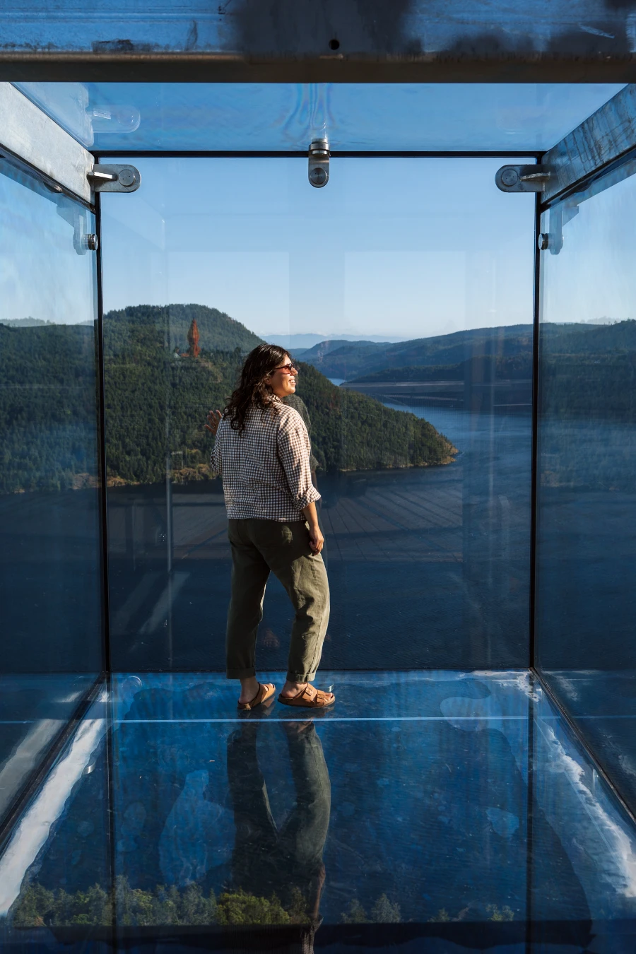 A woman stands in a glass cube at the top of the Malahat Skywalk with the Saannich Inlet below