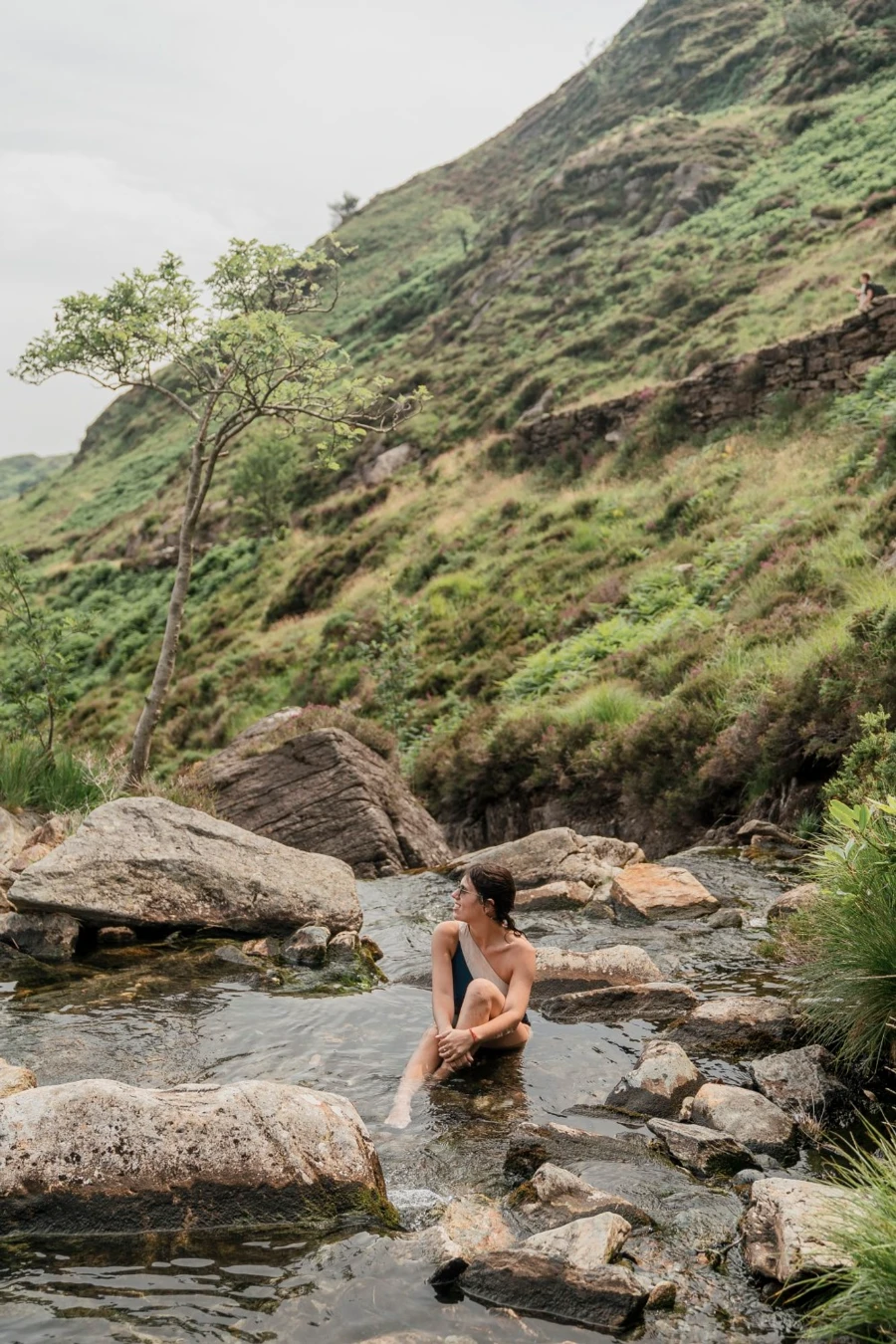 A woman sitting in a shallow pool of water with a large rolling green hill behind her
