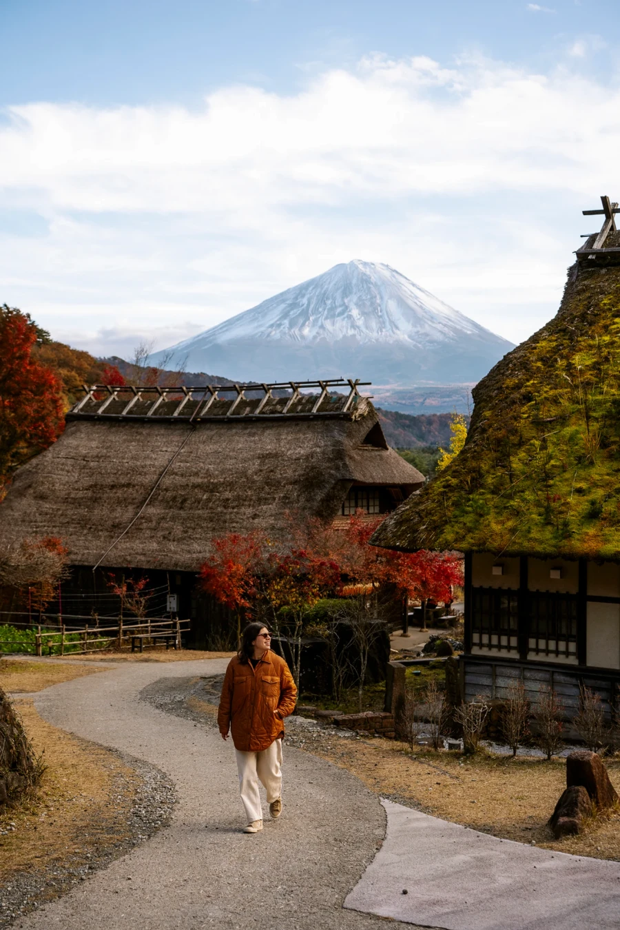 A woman walks along a gravel path past the thatched roof houses of  Saiko Iyashi-no-Sato Nenba