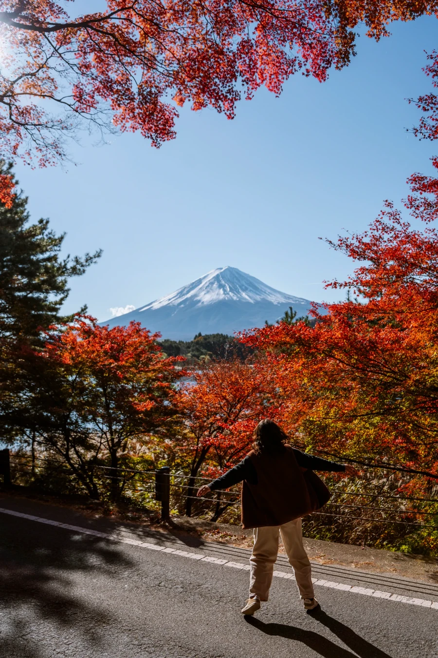 A woman spins in circles along a paved road that is lined with red bushes that perfectly frame Mt Fuji in the distance