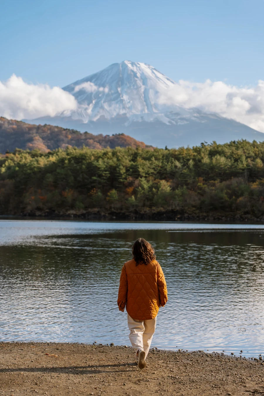 A woman walking along the sand towards the calm water of Saiko Lake with Mt Fuji in the disttance