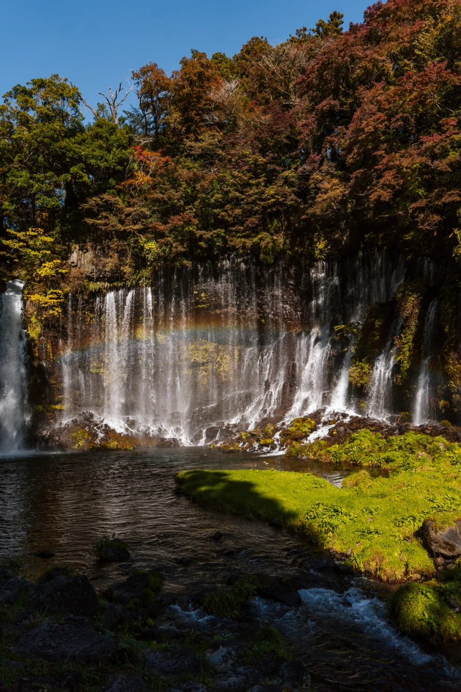 Several cascades of water fall into a mossy pool with their mist creating a rainbow at Shiraito Falls, Japan