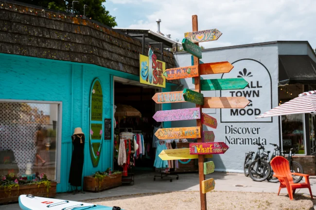A multicoloured signpost points out in many different directions at several random destinations around the world, with a large building in the background with a mural painted on the facade that reads ‘Small Town Big Mood Discover Naramata’