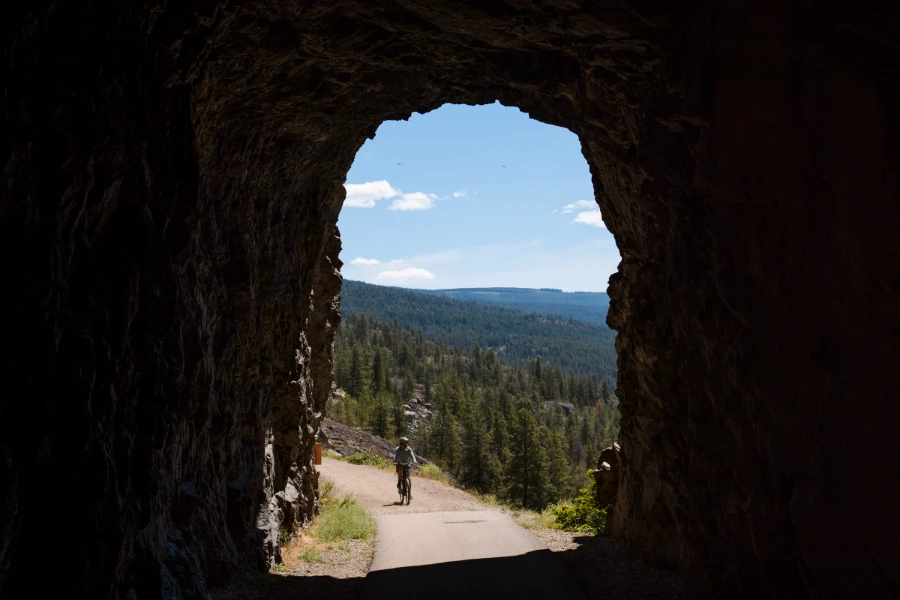 A cyclist rides along a wide gravel path entering the mouth of a large tunnel carved into the rock face