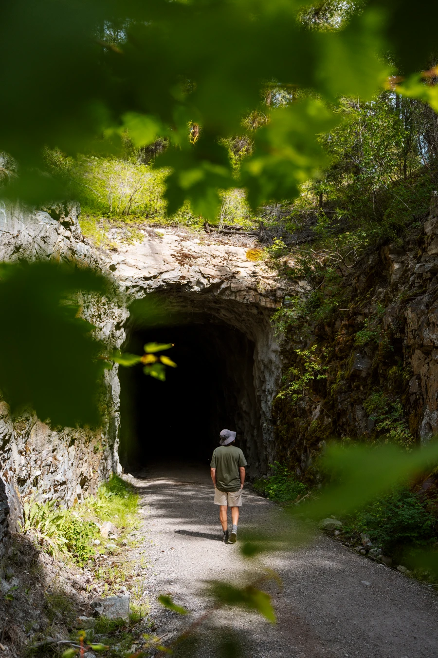 Framed by leaves of a tree in the foreground, a man walks along a flat and wide gravel path into a rectangularly cut tunnel.