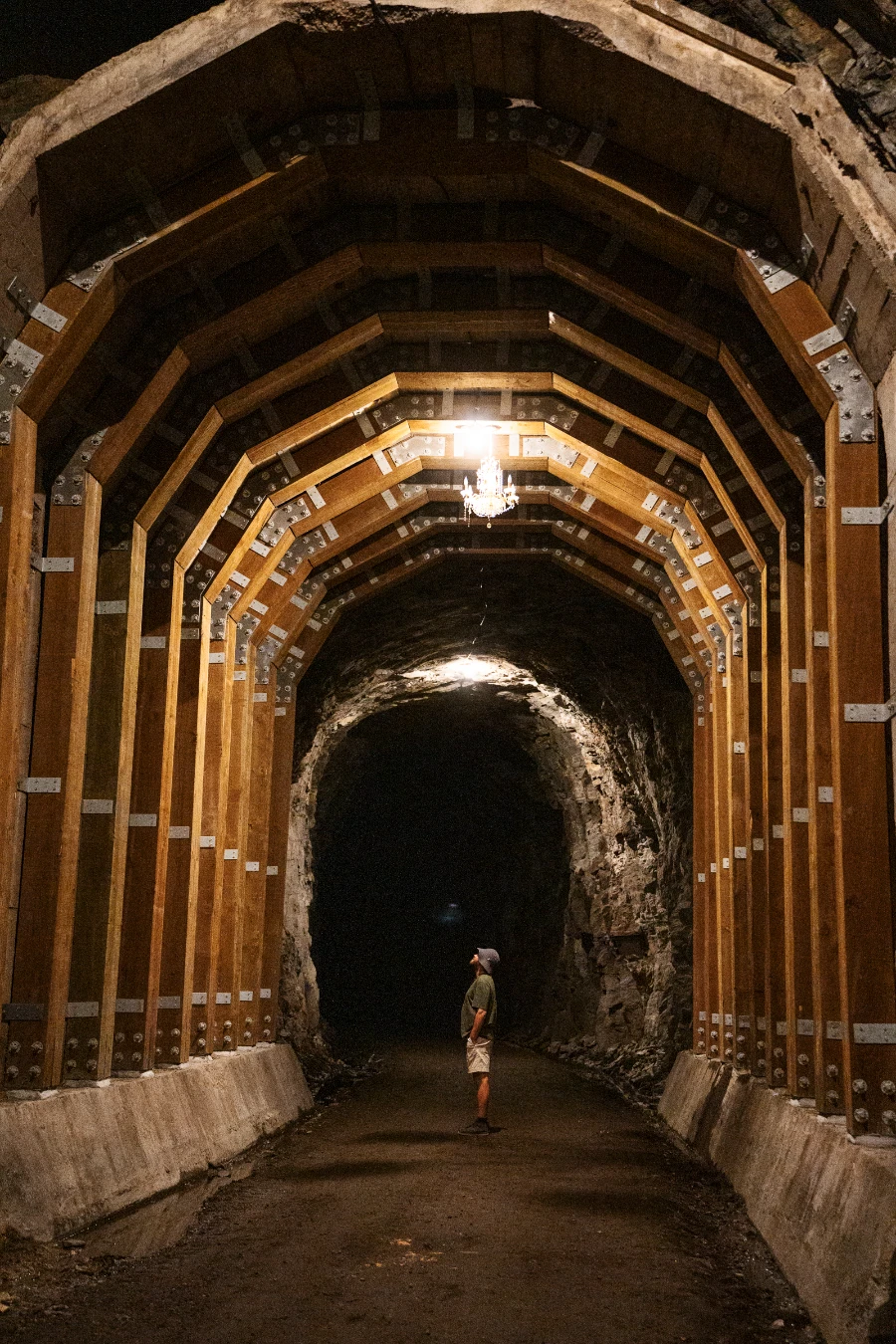 A man stands in a dark tunnel lit by a chandelier hanging from the ceiling and lined with large wooden reinforcements.