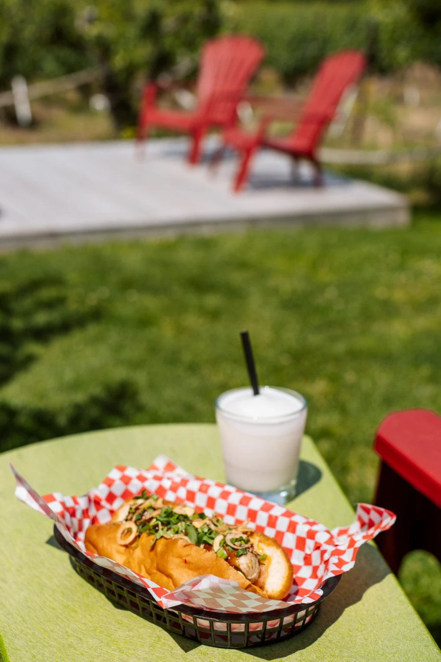 A hot dog overflowing with toppings in a paper basket next to a wine slushie with more red Muskoka chairs out of focus in the distance