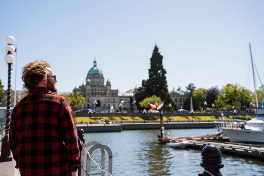 A man wearing a red flannel standing on the waterfront path in Victoria BC with the parliament building in the background