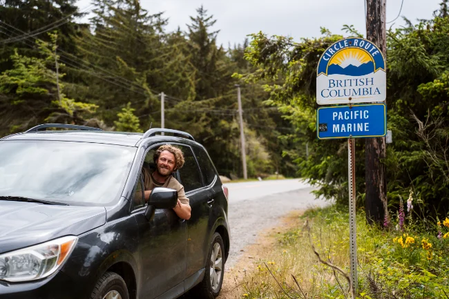 A man leans his head out of grey Subaru Forester parked on the side of the road next to a road sign that reads “Pacific Marine Circle Route”
