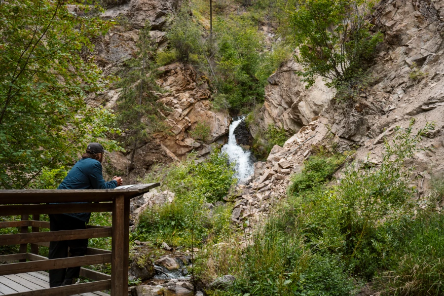 A man stands on a wooden platform and leans on a wooden fence looking out at a small waterfall
