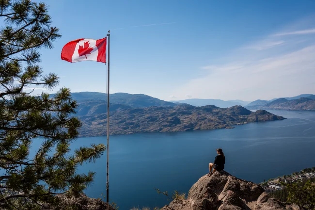 A woman sits on a boulder at the top of a mountain next to a waving Canadian flag and high above Okanagan Lake that stretches and turns and disappears beyond mountains in the distance