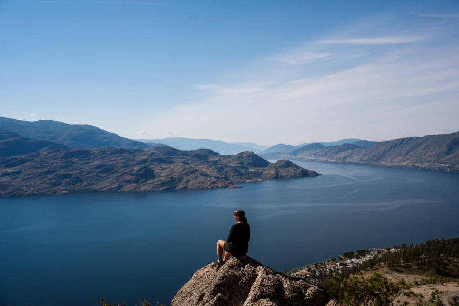 A woman sits on a boulder at the top of Pincushion Mountain looking down at the vast expanse of Okanagan Lake