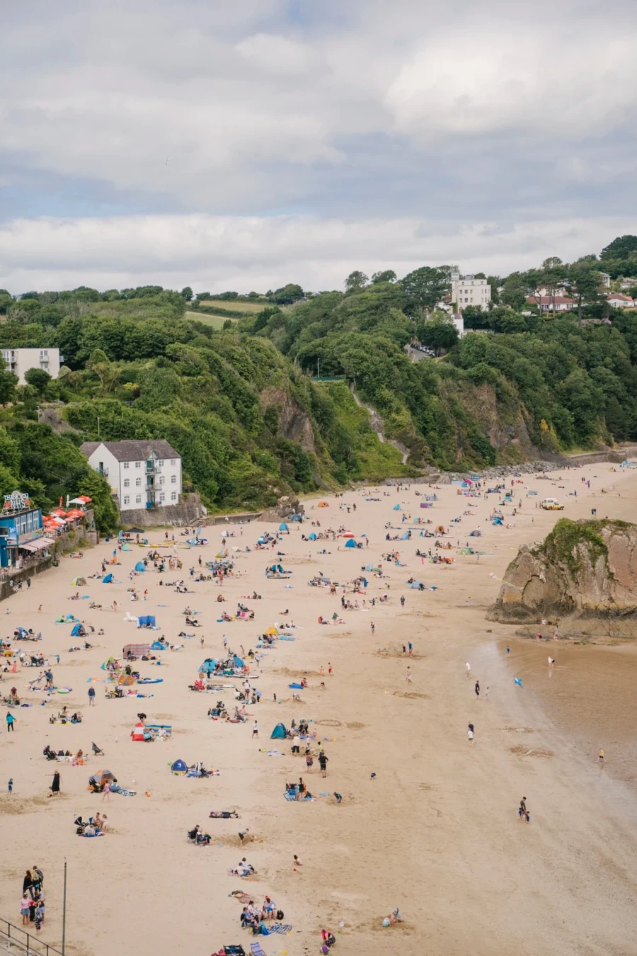 Crowds of people lounging on a white sand beach bordered by a forested cliff in Tenby, Wales