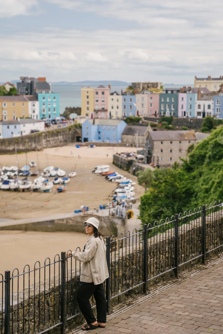 A woman leans on a black metal fence looking down at a white sand beach lined with colorful houses in Tenby, Wales