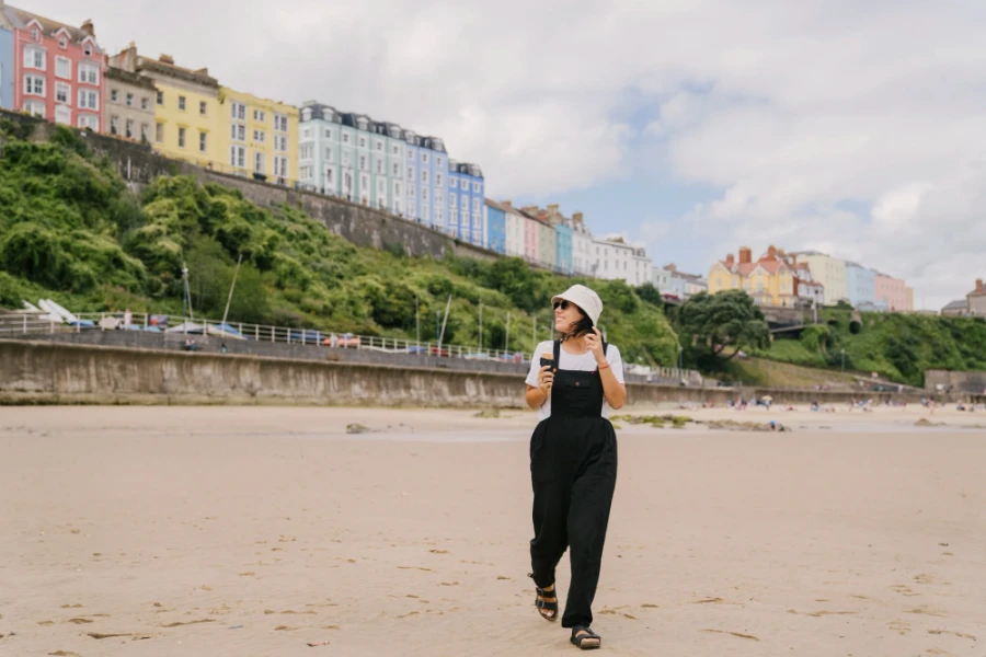 A woman eating an icrecream cone walks along a wet sandy beach with colorful houses behind her atTenby Beach