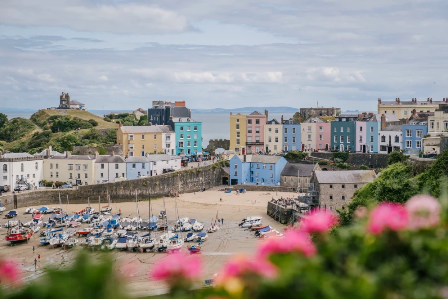 A row of pastel colored houses sit above a sandy beach with boats sitting on the sand in front of them at low tide in Tenby