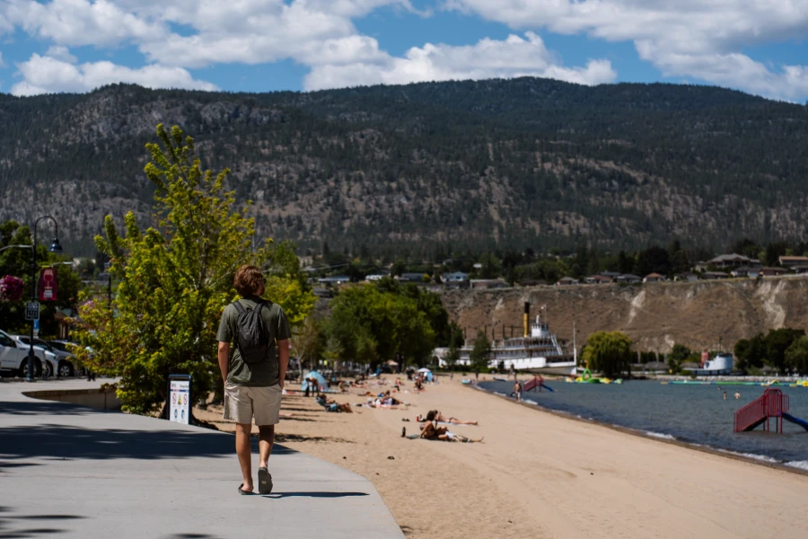 A man walks along a wide cement sidewalk in Penticton, looking out to his right at the wide sandy shores of Okanagan Lake Beach, where several people are lying out on towels in the sunshine, with the SS Sicamus visible at the far end of the beach.
