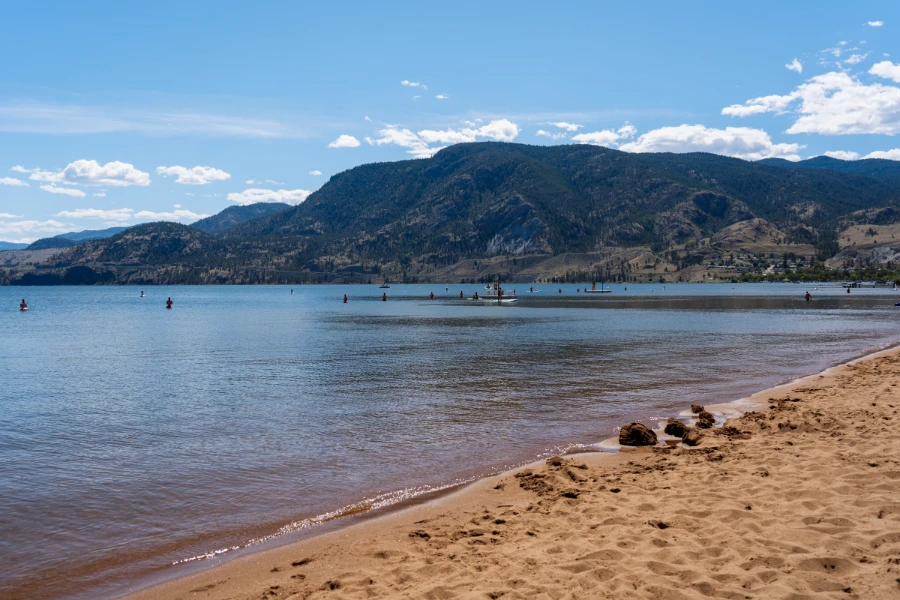 The sandy shores of Skaha Lake Beach next to a calm, wave-less lake with several swimmers and paddle boarders who look small in the distance, dwarfed further by the large mountain looming on the horizon.