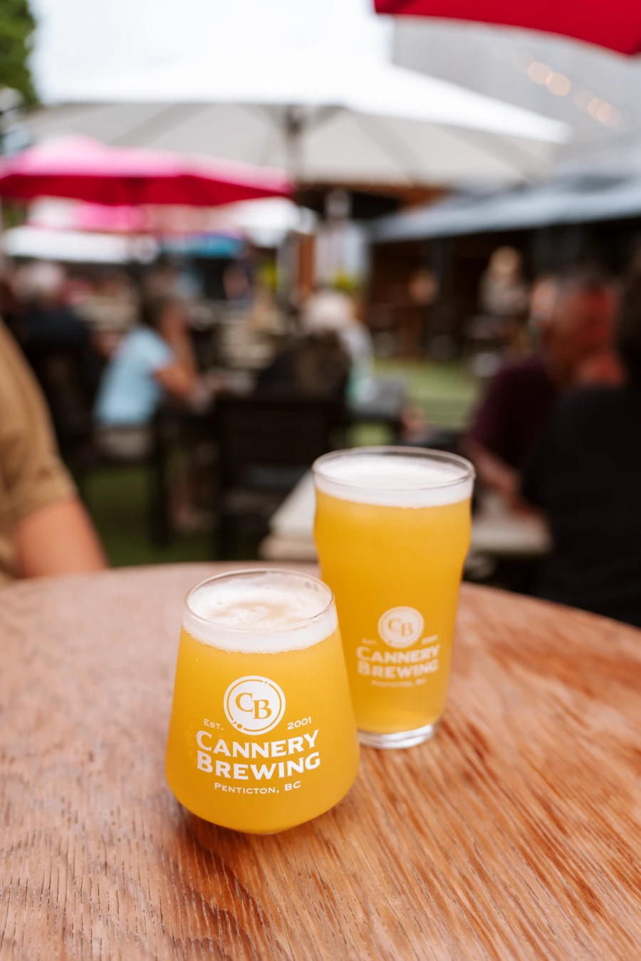 Two hazy beers in different-shaped glasses that both have the words ‘Cannery Brewing’ written on them sit on a wooden table with several other busy tables out of focus in the distance.
