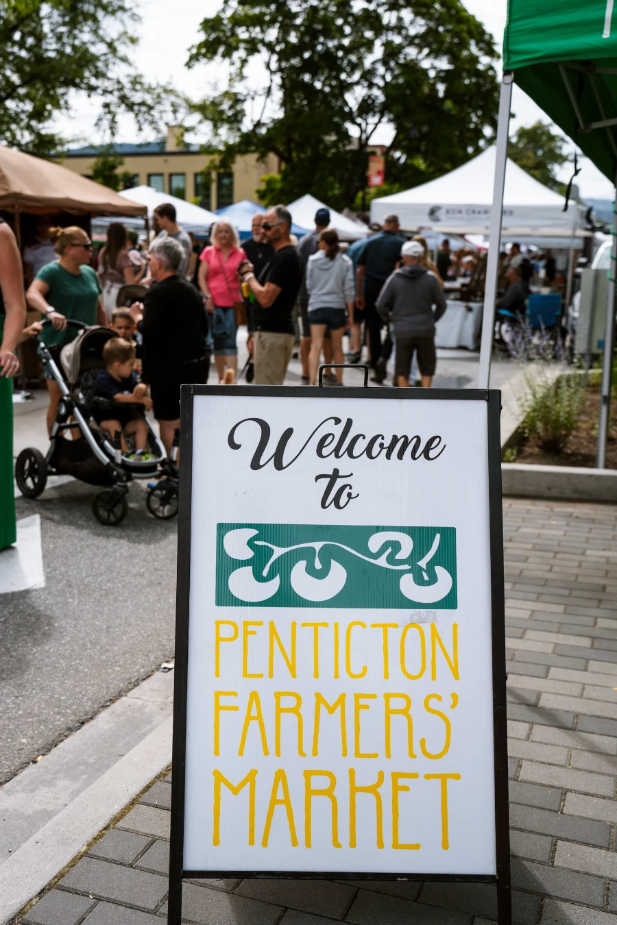 A sandwich board style sign reads ‘Welcome to Penticton Farmers’ Market’ with a large group of people behind it wandering through a row of several market booths.