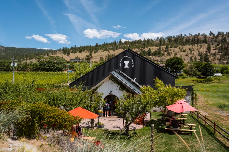 A grassy patio area with picnic tables, Muskoka chairs, and shade trees sits in front of an old farmhouse that has been converted into a modern brewery has a sign near the apex of the A-frame structure that reads ‘Abandoned Rail Brewing’