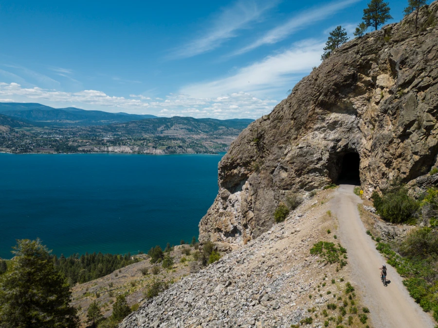 A woman riding an E-bike along the gravel KVR trail heading into a dark tunnel known as ‘Little Tunnel’ with the striking blue of Okanagan Lake far below the path to the left.