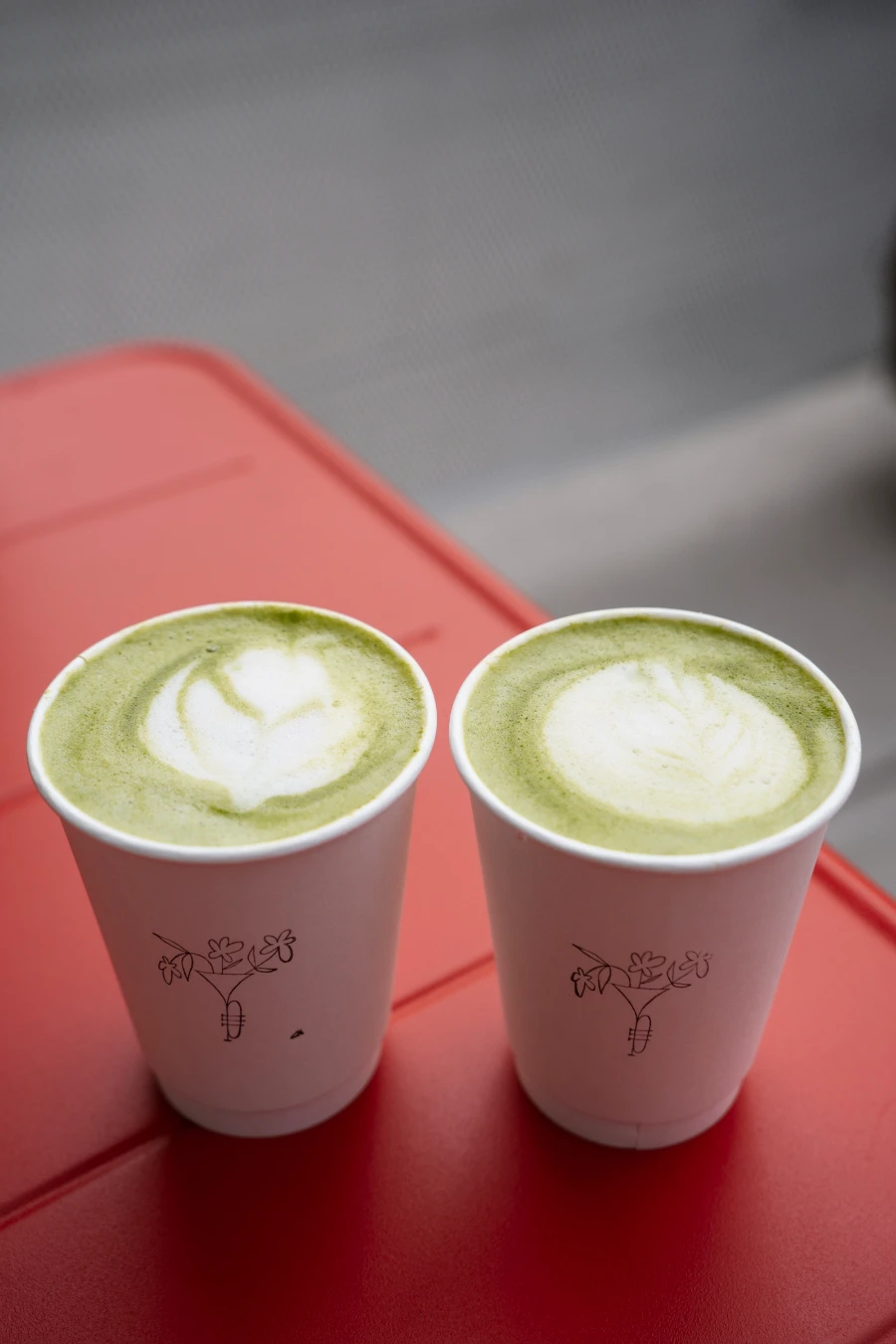 Two Matcha Lattes in paper cups sit on a red metal table at Soe Cafe in Penticton.