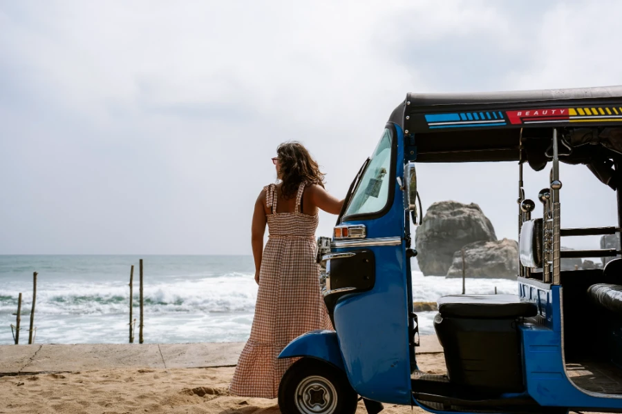 A woman stands beside tuktuk on the coast as huge white wash waves crash onto the shore