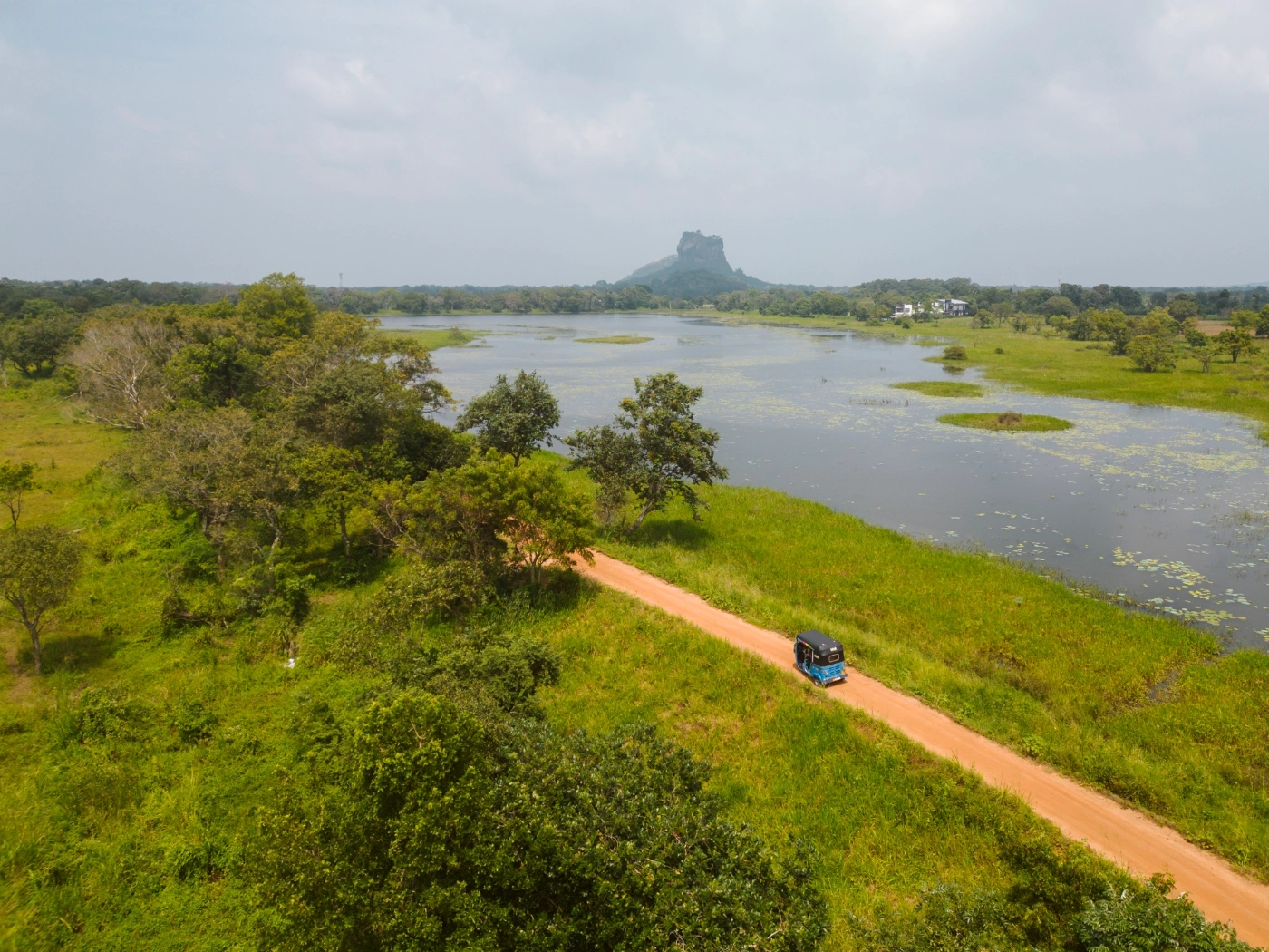 A blue tuktuk driving along a dirt road next to a lake with Lion's Rock in the distance in Sigiriya, Sri Lanka